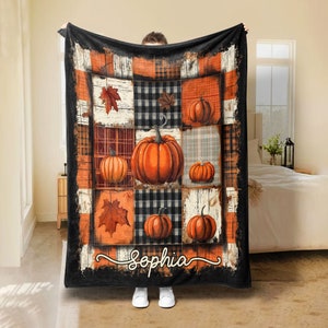 Peut inclure: Une couverture sur le thème de l'automne avec un motif patchwork de citrouilles, de feuilles et de motifs à carreaux orange, noir et blanc. La couverture porte le nom "Sophia" en écriture cursive.