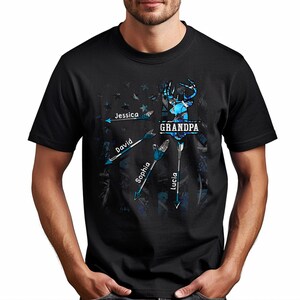 May include: Black t-shirt with a blue and white camouflage deer head design. The text "GRANDPA" is above the deer head. Four arrows point to the deer head with the names "Jessica", "David", "Sophia", and "Lucia" written on them.