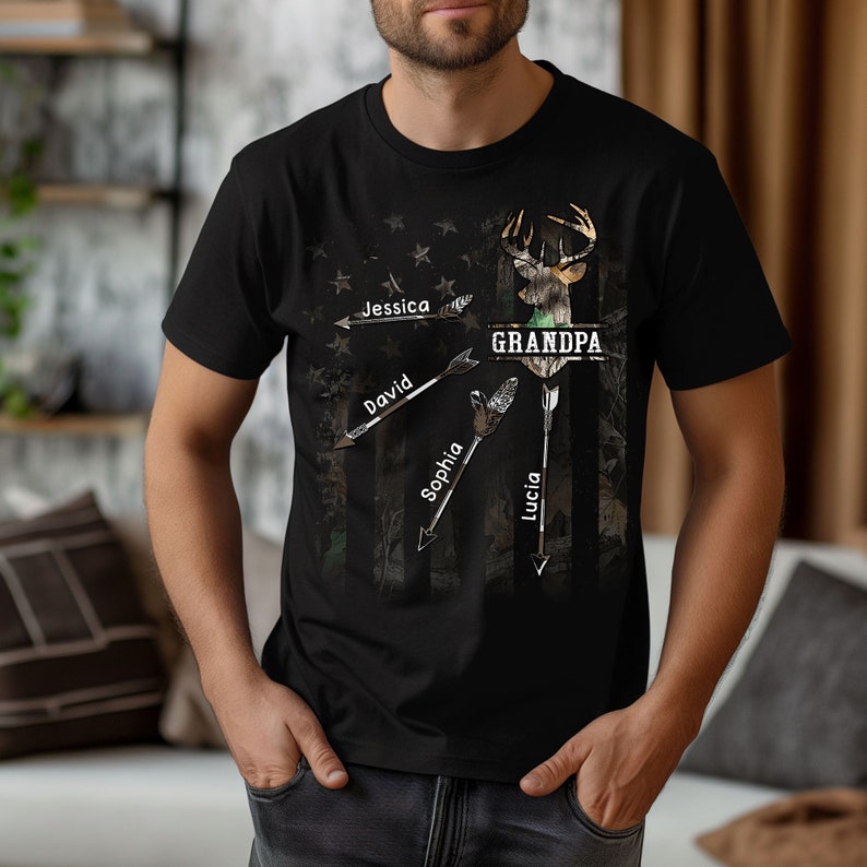 May include: Black t-shirt with a camouflage pattern and a deer silhouette. The text "GRANDPA" is printed above the deer. Four arrows point to the deer, each with a name written on it: Jessica, David, Sophia, and Lucia.