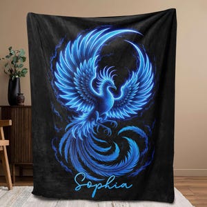 May include: A black blanket featuring a vibrant blue phoenix design. The phoenix is depicted with outstretched wings and a flowing tail, with the name "Sophia" written in cursive script at the bottom. The design has a glowing effect.