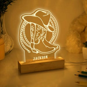 May include: A wooden night light with a white acrylic insert featuring a cowboy boot and hat design. The name "JACKSON" is printed on the base of the light.