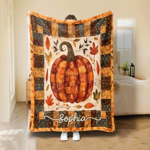 May include: A cosy autumn-themed blanket with a large orange pumpkin illustration. The blanket has a cream background with autumnal leaves and the name "Sophia" in cursive. The border features a patchwork design with orange, brown, and navy blue squares.