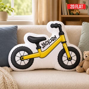 May include: A white shaped throw pillow featuring a yellow balance bike illustration with black tires and handlebars. The name JACKSON is printed in black script on the frame. The pillow has a die-cut silhouette matching the bike shape.
