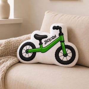May include: A white throw pillow shaped like a balance bike with a green frame, black wheels, and a black seat. The word "JACKSON" is printed in black lettering on the frame of the bike. The pillow features a detailed graphic illustration.