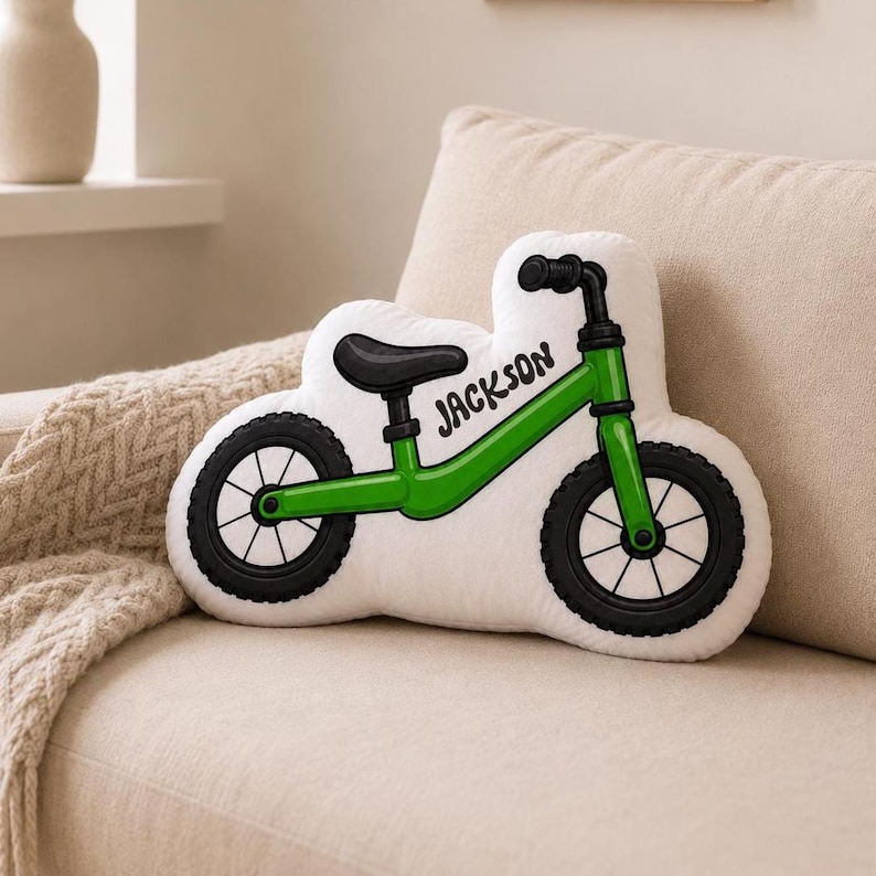 May include: A white throw pillow shaped like a balance bike with a green frame, black wheels, and a black seat. The word "JACKSON" is printed in black lettering on the frame of the bike. The pillow features a detailed graphic illustration.