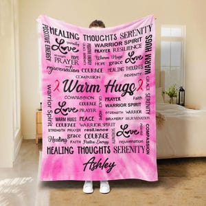 May include: Pink and white fleece blanket with a pink ribbon and the words "Warm Hugs" in black. The blanket is covered in words of encouragement and support, including "Love", "Hope", "Courage", "Warrior Spirit", "Healing", "Prayer", "Resilience", "Serenity", and "Compassion". The name "Ashley" is printed at the bottom of the blanket.