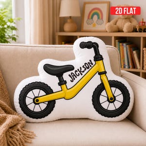 May include: A white throw pillow shaped like a balance bike with a yellow frame, black seat, black handlebars, and black tires. The name JACKSON is printed in black script on the yellow frame. The pillow features a 2D flat graphic design.