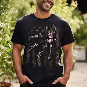 May include: Black t-shirt with a camouflage pattern and a pink deer silhouette. The text "GRANDPA" is printed in pink above the deer. Four arrows point to the deer, each with a name printed on it: Jessica, David, Sophia, and Lucia.