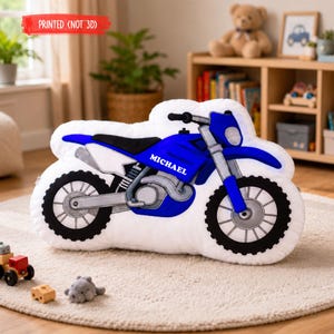Puede incluir: Un coj&iacute;n blanco con forma de motocicleta con un dise&ntilde;o azul y negro. El nombre "MICHAEL" est&aacute; impreso en el lateral. El texto "PRINTED (NOT 3D)" est&aacute; impreso en una pancarta roja.