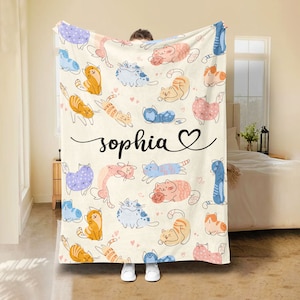 Op de afbeelding: Een zachte, crèmekleurige fleece deken met een herhalend patroon van kleurrijke cartoon katten. De deken heeft de naam "sophia" in zwarte cursieve letters met een hartje.