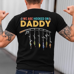 May include: Black t-shirt with a fishing rod graphic and the text "We are hooked on Daddy".  The fishing line has six hooks with names written on them.