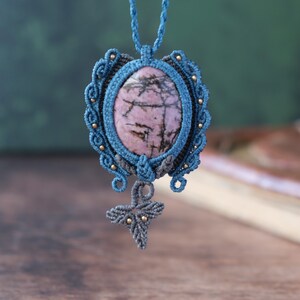 Op de afbeelding: Een blauwe en grijze macramé ketting met een roze en zwarte edelsteen hanger. De hanger is ovaalvormig en heeft een geknoopt ontwerp rond de rand. De ketting heeft een kleine, geknoopte vlinderhanger die aan de onderkant hangt.