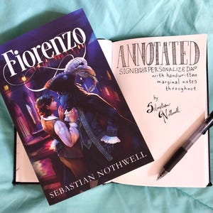 May include: A hardcover book titled "Fiorenzo" with a fantasy illustration of two figures in a dark setting. An open notebook displays handwritten text: "ANNOTATED SIGNED & PERSONALIZED..." A black pen rests on the notebook.