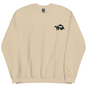 May include: A beige sweatshirt with a crew neck and long sleeves. A small embroidered badger in black and white is on the upper left chest. The Gildan brand tag is visible on the collar.