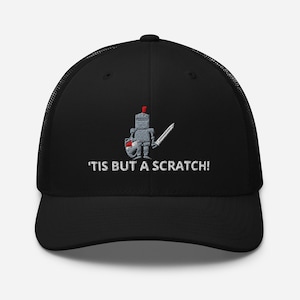 May include: Black trucker cap with a mesh back and a curved peak. The front displays an embroidered cartoon knight with a sword and shield, and the text "'TIS BUT A SCRATCH!"