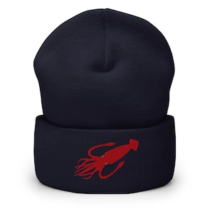 Puede incluir: Un gorro azul marino con un diseño de calamar rojo bordado en el borde doblado. El diseño del calamar presenta tentáculos y dos ojos amarillos. El gorro está hecho de un material de punto suave.