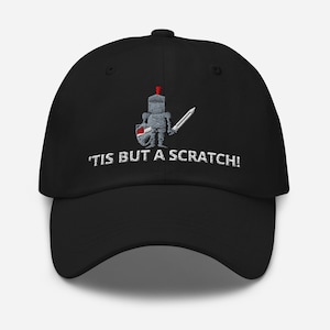 May include: Black baseball cap with a white embroidered design. The design features a cartoon knight holding a sword and shield, with the text "'TIS BUT A SCRATCH!" in white.