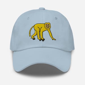 Può includere: Cappellino da baseball azzurro chiaro con un motivo a scimmia ricamato in giallo. La scimmia è raffigurata mentre cammina, con piedi neri e un viso dettagliato. Il cappellino ha una visiera curva e un cinturino regolabile.