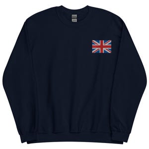 Puede incluir: Sudadera azul marino con una pequeña bandera de la Union Jack bordada en el pecho izquierdo. La bandera es roja, blanca y azul. La sudadera es de color liso y tiene cuello redondo.
