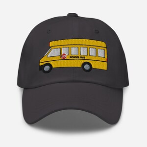 May include: A dark grey baseball cap with an embroidered yellow school bus design. The school bus has black wheels, a red stop sign, and the words "SCHOOL BUS". The cap has a curved peak and an adjustable strap.
