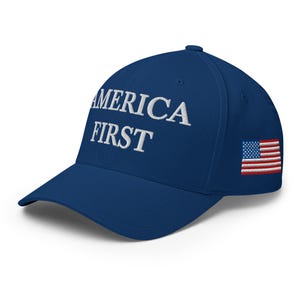 May include: A navy blue baseball cap with the embroidered text "AMERICA FIRST" in white. The cap features a small embroidered American flag on the side. The hat has a curved brim and a structured fit.