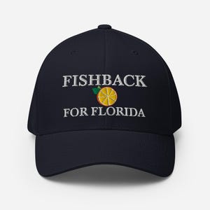 May include: A navy blue baseball cap with the text "FISHBACK FOR FLORIDA" embroidered in white. A yellow and orange citrus fruit graphic is centered above the text. The cap is a fitted style.