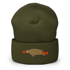 Puede incluir: Gorro verde oliva con un diseño bordado de ornitorrinco. El ornitorrinco es marrón con pico y cola naranjas. Un accesorio cálido y cómodo.
