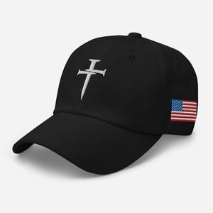 May include: Black baseball cap with a white embroidered cross and nail design on the front. The cap features the American flag on the side. A stylish and comfortable accessory.