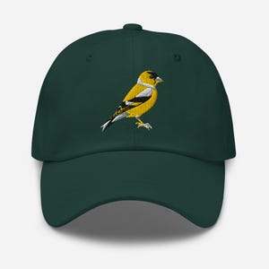 Könnte beinhalten: Dunkelgrüne Baseballkappe mit einem gestickten Stieglitz. Der Vogel hat gelbe, schwarze und weiße Details. Die Kappe hat einen gebogenen Schirm und einen Knopf oben.