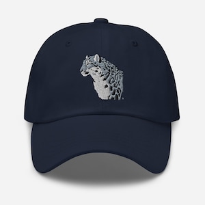 May include: A navy blue baseball cap with an embroidered snow leopard design. The cap has a curved peak and an adjustable strap. The snow leopard is rendered in white and grey thread, with detailed spots and facial features.