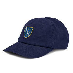 May include: A navy blue corduroy baseball cap with the Bosnian coat of arms embroidered on the front. The emblem is a yellow shield with a white stripe and blue fleur-de-lis symbols. A classic, adjustable hat.