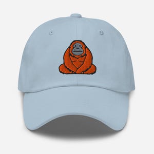 Könnte beinhalten: Hellblaue Baseballkappe mit einem gestickten Orang-Utan-Design. Der Orang-Utan ist in Orange- und Brauntönen mit einem grauen Gesicht dargestellt. Die Kappe hat einen gebogenen Schirm und einen verstellbaren Riemen am Rücken.