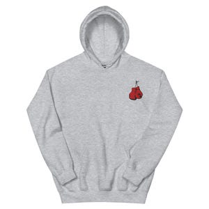 May include: A light gray hoodie with a drawstring hood and a kangaroo pocket. The hoodie features a small embroidered design of two red boxing gloves on the upper left chest area. The sweatshirt is made of a soft material.