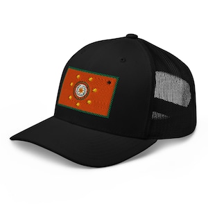 Puede incluir: Gorra de camionero negra con un parche rectangular con fondo naranja, un diseño circular central y detalles en forma de estrella. El parche está delineado en verde. La gorra tiene una parte trasera de malla.