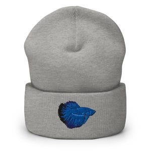 May include: A gray beanie with a folded brim. The beanie features an embroidered blue betta fish design on the front. The fish has detailed fins and scales, adding a touch of aquatic flair to the winter accessory.