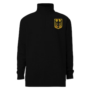 Könnte beinhalten: Schwarzes Sweatshirt mit Viertelreißverschluss und hohem Kragen. Das Sweatshirt hat ein goldfarbenes, schwarzes und rotes Emblem eines Adlers auf der oberen linken Brust. Das Emblem befindet sich auf einem goldenen Schild. Das Sweatshirt hat lange Ärmel.