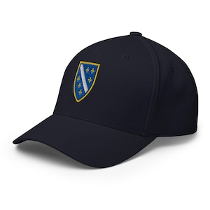 May include: A navy blue baseball cap featuring an embroidered emblem. The emblem is a shield shape with a blue background, a white diagonal stripe, and yellow fleur-de-lis symbols. The cap has a curved brim and a structured crown.