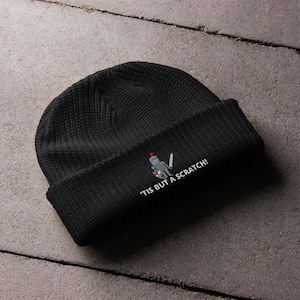 May include: Black knitted beanie featuring an embroidered design of a knight with a sword and shield. The text "'Tis but a scratch!" is printed below the design in white.