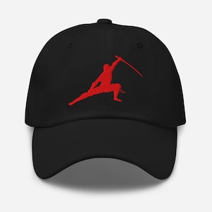 May include: Black baseball cap featuring a red silhouette of a person in a martial arts stance, holding a curved sword. The cap has a curved brim and a button on top. The design is embroidered on the front.
