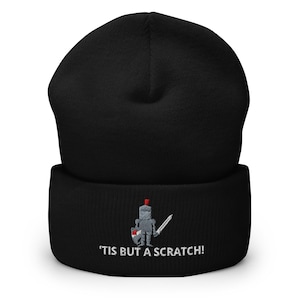 May include: Black beanie with an embroidered design of a knight holding a sword and shield. The text "'TIS BUT A SCRATCH!" is below the design. The beanie is made of a soft, knitted material.