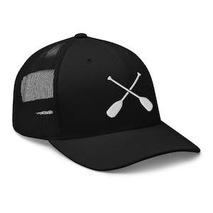 Puede incluir: Gorra de camionero negra con un diseño bordado blanco de dos remos cruzados. La gorra tiene una parte trasera de malla y una visera curva. Un accesorio elegante para los entusiastas del aire libre.