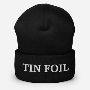 以下が含まれることがあります： 白い「TIN FOIL」の刺繍が入った黒いビーニー。ビーニーは柔らかいニット素材でできており、折り返しのあるつばが付いています。帽子は無地の黒色です。