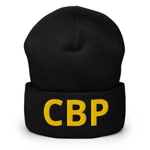 Könnte beinhalten: Schwarze Mütze mit den gelb gestickten Buchstaben "CBP". Die Mütze ist aus einem weichen Material gefertigt und hat einen gefalteten Saum. Ein warmes Accessoire für kaltes Wetter.