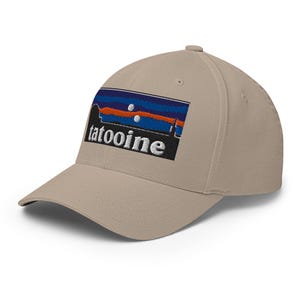 Puede incluir: Gorra de béisbol beige con visera curva. La gorra presenta un parche bordado con la palabra "tatooine" en blanco, debajo de un diseño estilizado en azul, naranja y negro.