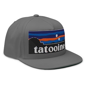 Puede incluir: Gorra gris de ala plana con un parche rectangular bordado. El parche presenta un paisaje estilizado con detalles azules, naranjas y blancos, y la palabra "tatooine" en letras blancas.