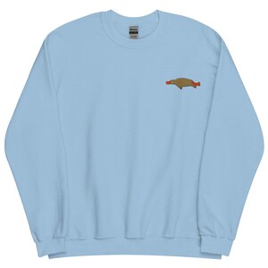 Puede incluir: Sudadera azul claro con un pequeño diseño bordado de un ornitorrinco marrón con pico y cola rojos en la parte superior izquierda del pecho. La sudadera tiene cuello redondo y mangas largas.