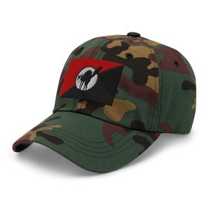 May include: Camouflage baseball cap with a black and red patch featuring a black cat silhouette within a circle. The cap is green, brown, and tan with a curved brim and a button on top. A stylish accessory.