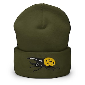 Puede incluir: Gorro verde oliva con un diseño de escarabajo bordado. El escarabajo es negro con un patrón de puntos amarillos. Este gorro de punto cálido y suave es ideal para el invierno.