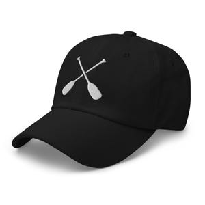 May include: Black baseball cap with a curved peak. The cap has a white embroidered design of two crossed oars. The hat is made of a soft material and has an adjustable strap at the back.