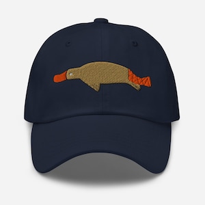 Puede incluir: Gorra de béisbol azul marino con un diseño bordado de ornitorrinco. El ornitorrinco tiene un cuerpo marrón, un pico naranja y una cola naranja. La gorra es un diseño clásico de seis paneles con una visera curva y una correa ajustable.